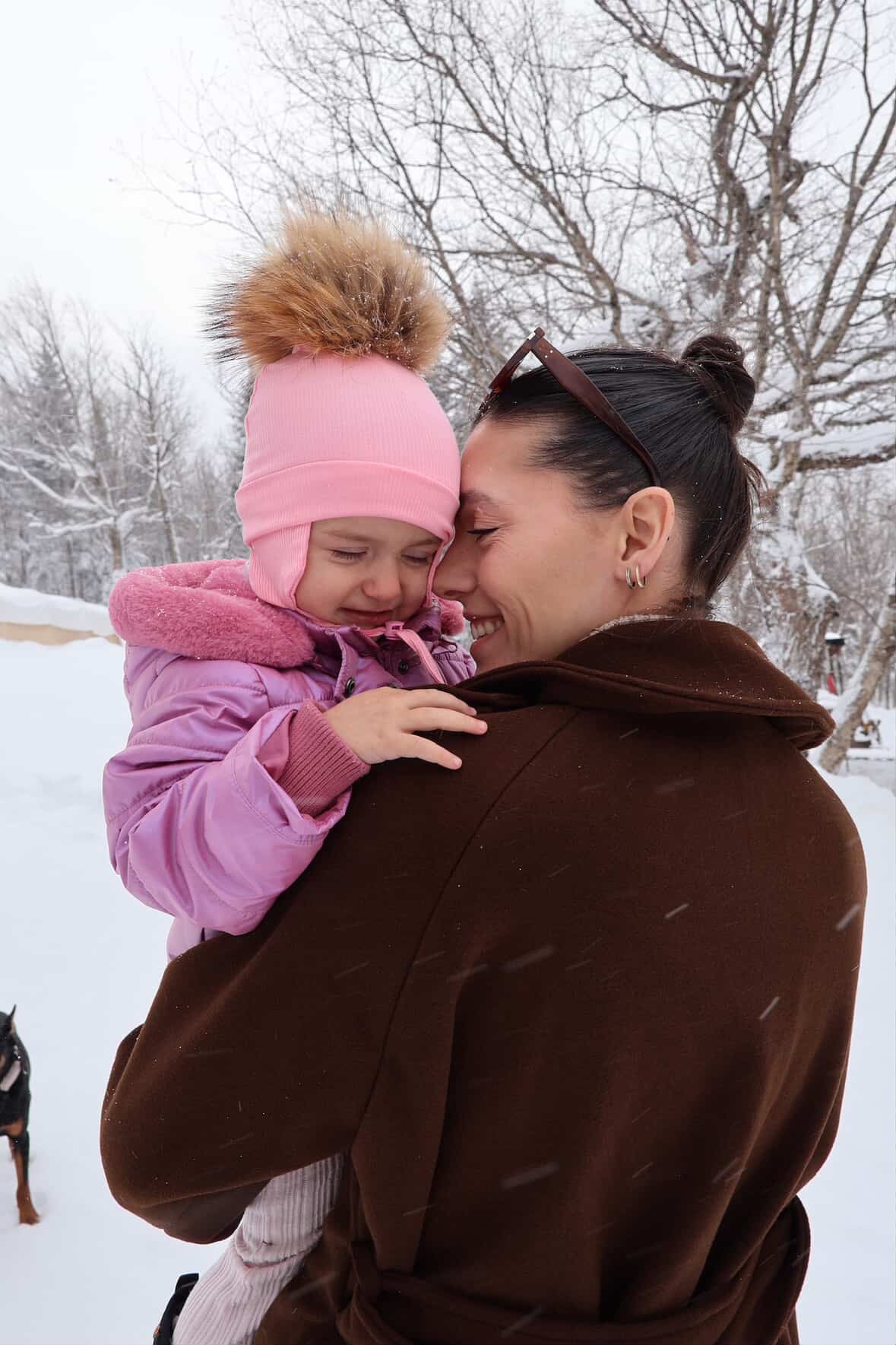 Bébé portant une tuque rose Kplla avec pompon dans les bras de sa maman dans la neige
